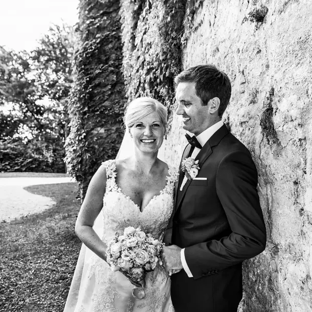 Hochzeitsfotografie, Paarshooting im Schlosspark von Schloss Langenburg. Brautpaar lehnt an der Mauer und Braut schaut lachend in die Kamera. Bräutigam schaut die Braut an.